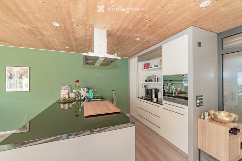 Modern kitchen with white cabinets, green walls, wooden ceiling, and stainless steel appliances. Features a central island with a black countertop, sink, and cutting board. Shelves hold various kitchen items, and a small table displays a decorative bowl a