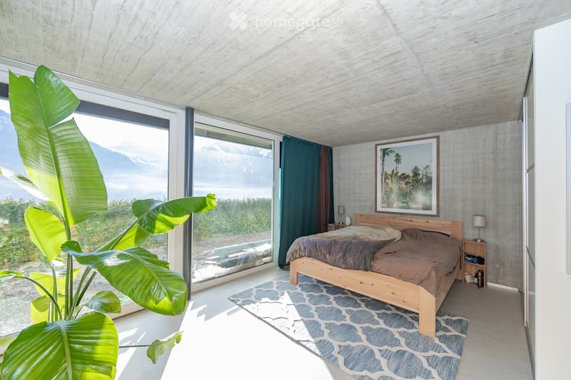 Modern bedroom with large windows offering a scenic mountain view, wooden bed, nightstands, and a large leafy plant near the window.