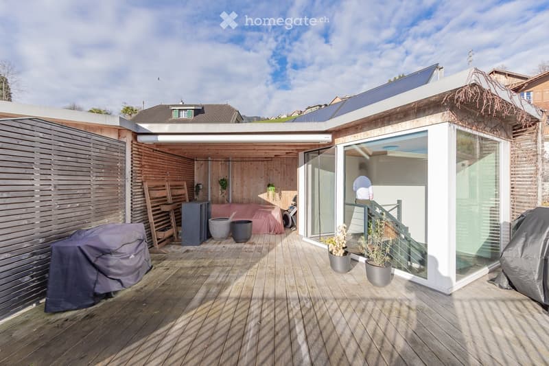 Wooden deck area with covered seating, potted plants, covered grill, and modern glass-walled structure.
