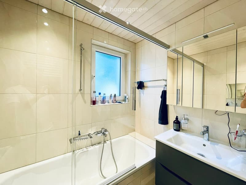 Modern bathroom with a bathtub, sink, mirror, and shower area. Features include a window, tiled walls, and various toiletries on the shelf.