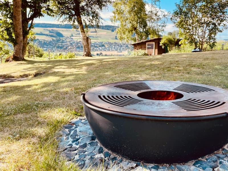 A modern outdoor fire pit with a sleek, metallic design is placed on a bed of stones in a grassy area, surrounded by trees and a scenic valley view.