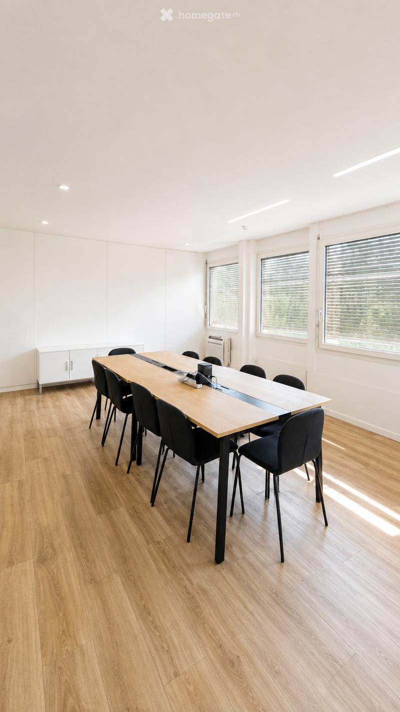 Bright, modern meeting room with a long wooden table surrounded by black chairs, a white cabinet, and large windows with blinds.