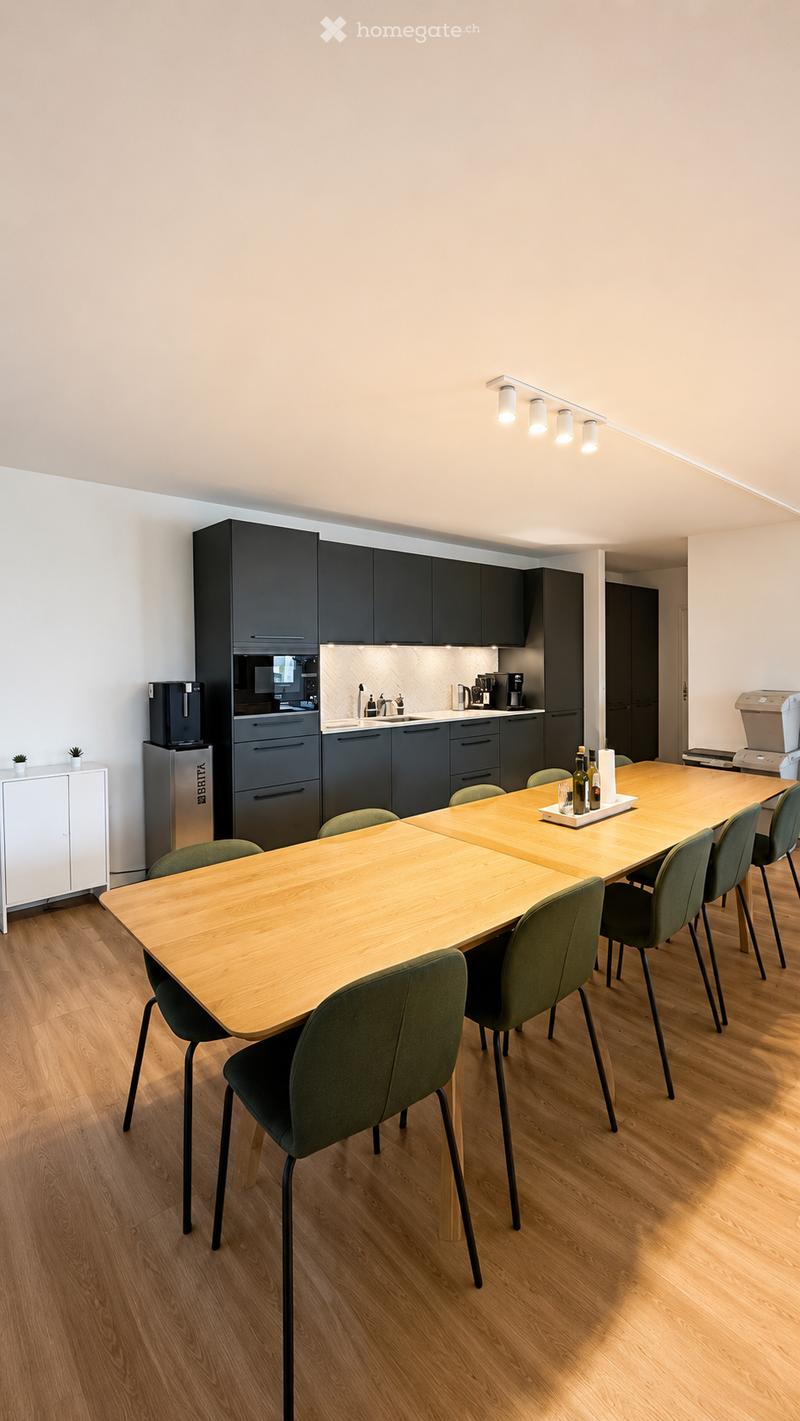 Modern kitchen and dining area with dark cabinets, a long wooden dining table, green chairs, and various appliances including a coffee machine and a printer.