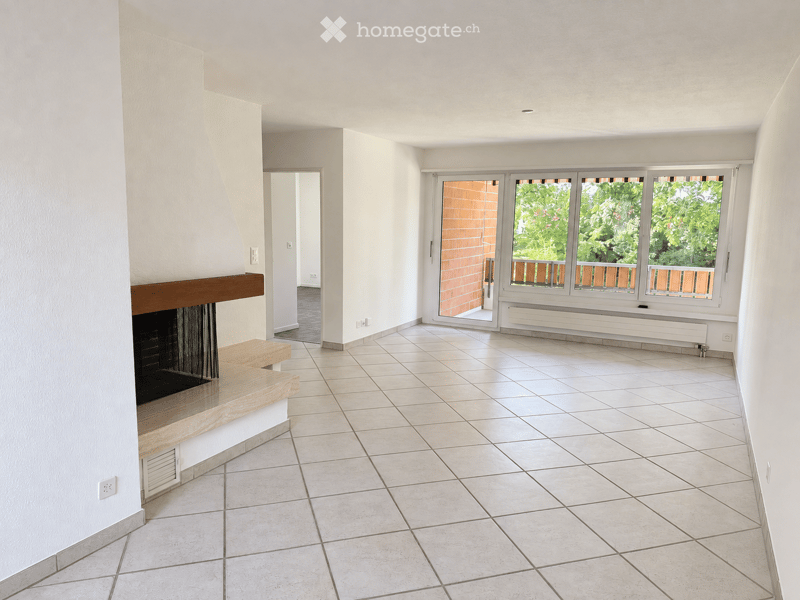 Empty room with tiled floor, white walls, fireplace with wooden mantel, large windows with wooden frames, and a sliding glass door leading to a balcony.
