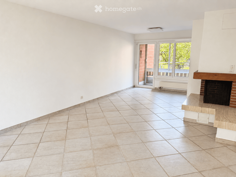 Empty room with tiled floor, white walls, fireplace, and large windows with a view of greenery outside.