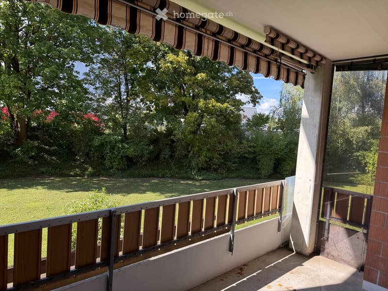 Balcony with wooden railing, striped awning, and view of a green lawn and trees.