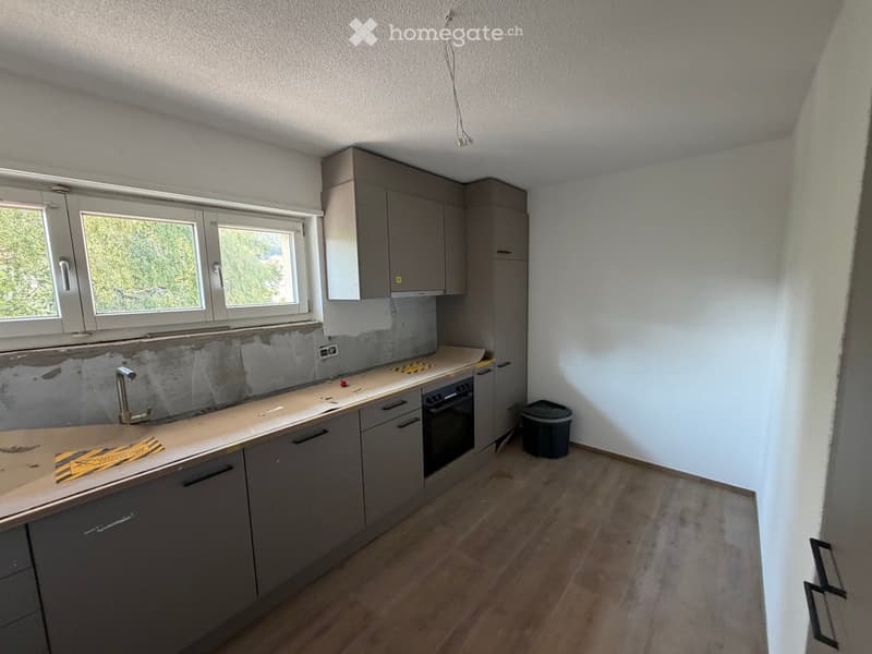 Empty kitchen with grey cabinets, a countertop, and a sink. The backsplash is partially removed, revealing the wall underneath.