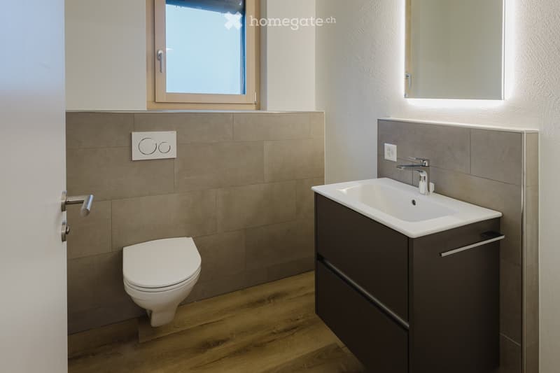 A compact bathroom with white sanitary fixtures, modern sink with a faucet, and a white toilet. The walls and floor have a warm, wooden texture, and there is a window that lets in natural light.