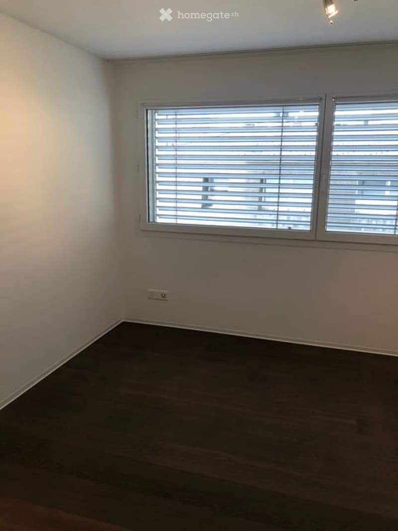 Empty room with wooden flooring, white walls, and a large window with blinds.