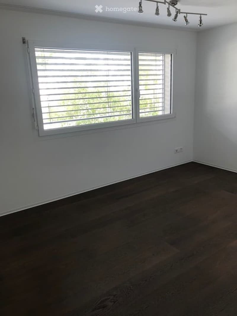 Empty room with wooden flooring, white walls, large window with blinds, and track lighting fixture.