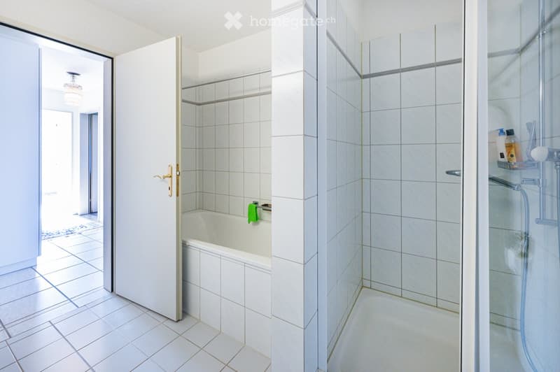 Modern bathroom with white tiled walls and floor, featuring a bathtub, glass-enclosed shower, and a mirror above the sink. A small shelf holds toiletries.