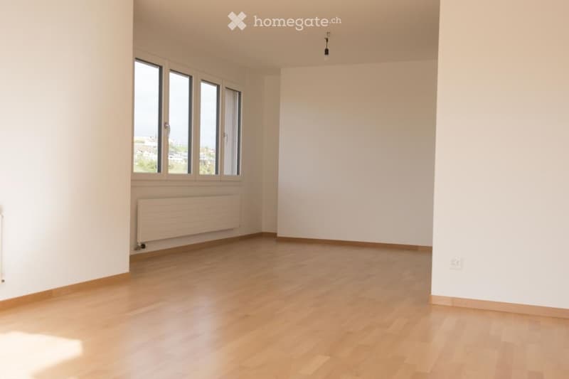 Empty room with light wooden flooring, white walls, large windows, and a radiator. No furniture or equipment present.
