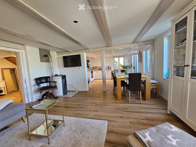 A modern, open-concept living and dining area with wooden flooring, a grey sofa, a glass coffee table, a dining table with chairs, a television mounted on the wall, and a small kitchen area visible in the background.