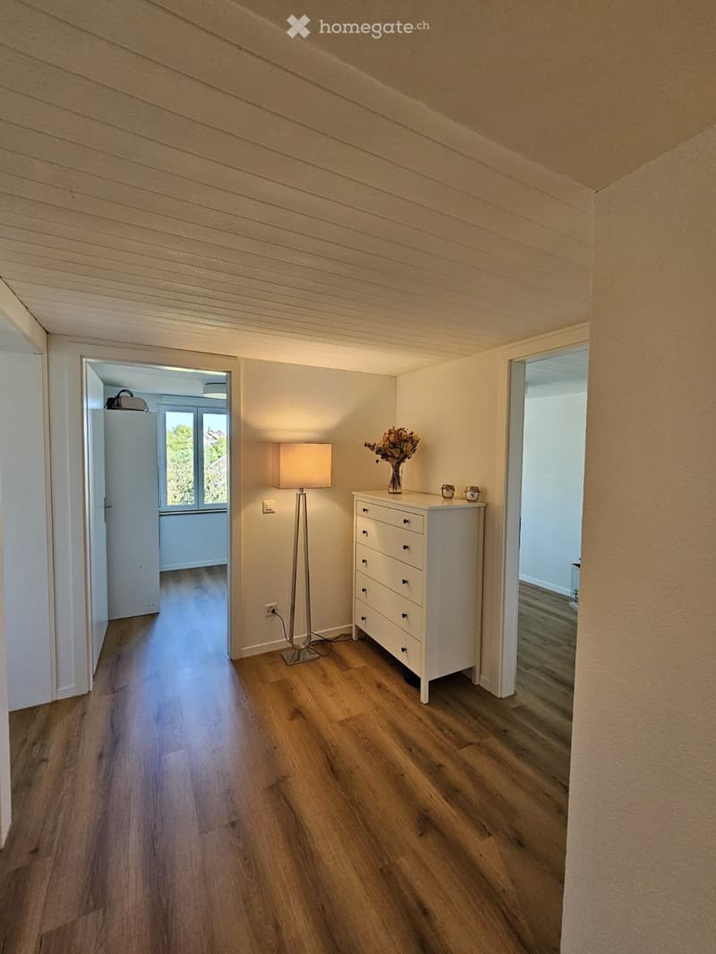 A narrow hallway with wooden flooring, white walls, and a slanted ceiling. A small white drawer unit with a vase of flowers sits near a tall table lamp. The hallway leads to an open area with large windows and a view of greenery.