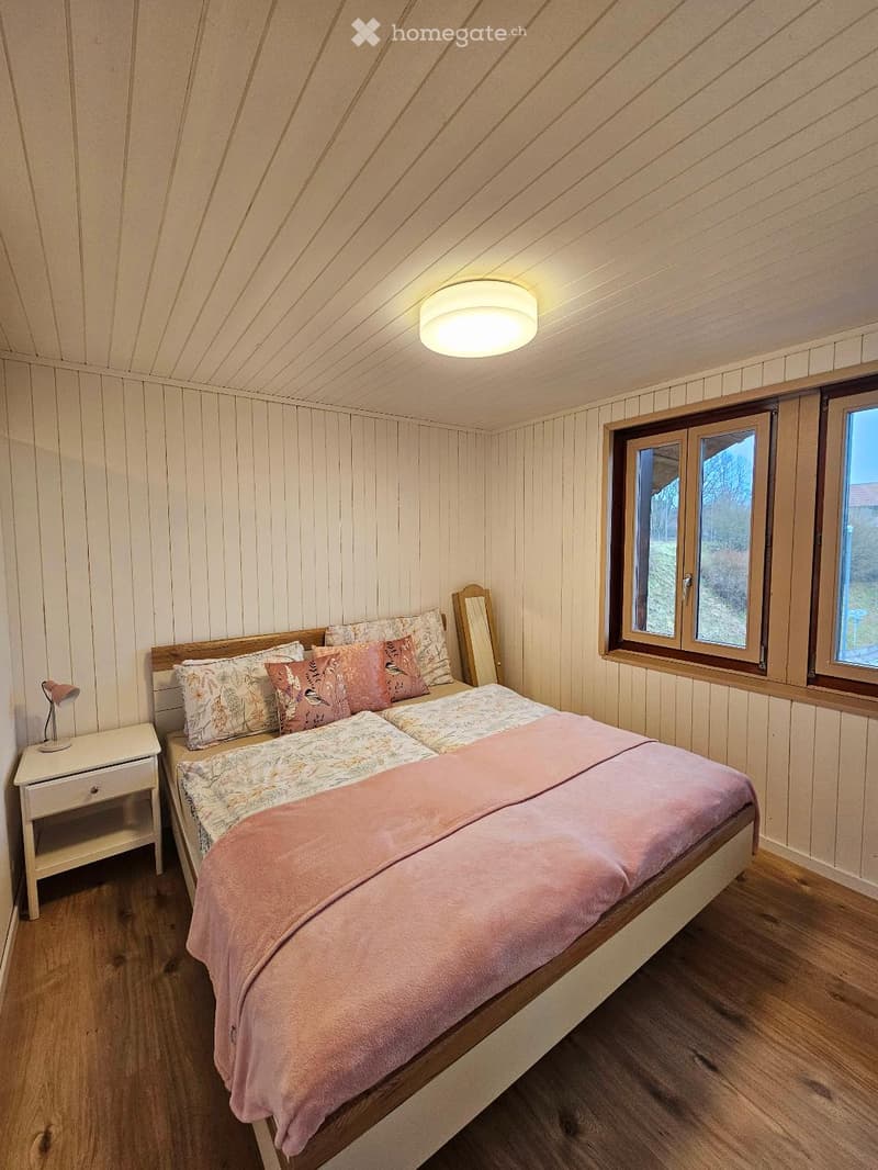 Cozy bedroom with wooden walls and ceiling, featuring a double bed with pink bedding, a nightstand with a lamp, and a mirror leaning against the wall. Large windows offer natural light.