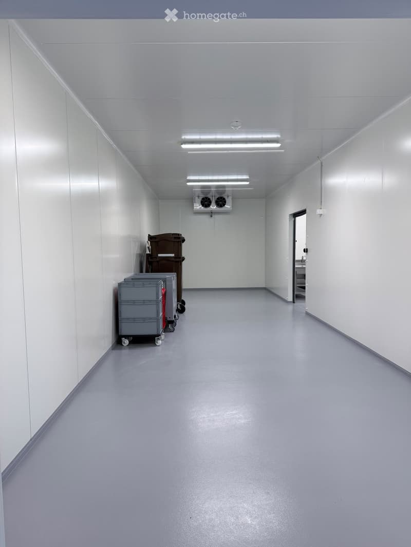 Clean, empty corridor with white walls, grey floor, fluorescent lighting, and a ventilation unit mounted on the wall.