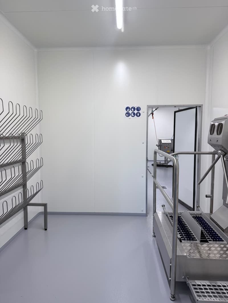 Empty room with white walls, grey floor, metal racks, and a stainless steel door leading to another area.