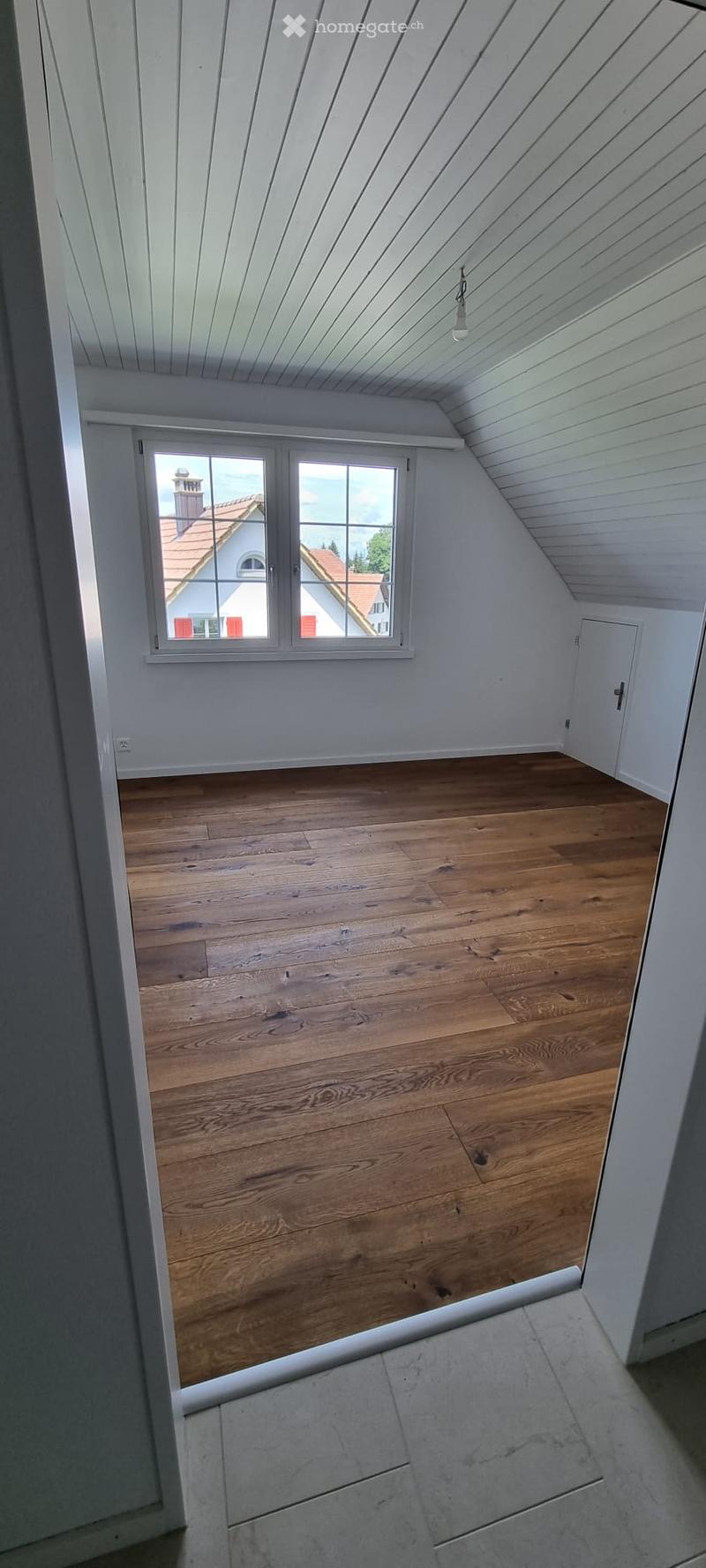 Empty attic room with wooden flooring, white walls, and a large window offering a view of the outside. The ceiling has a sloped design with exposed wooden beams.