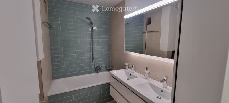 Modern bathroom with blue tiled walls, a bathtub, double sink vanity, and a large mirror with built-in lighting.
