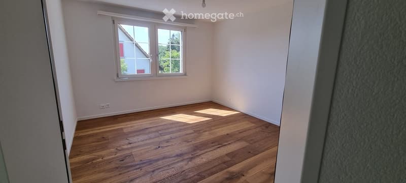 Empty room with wooden flooring and white walls, featuring a large window with white frame and blinds, allowing natural light to enter.