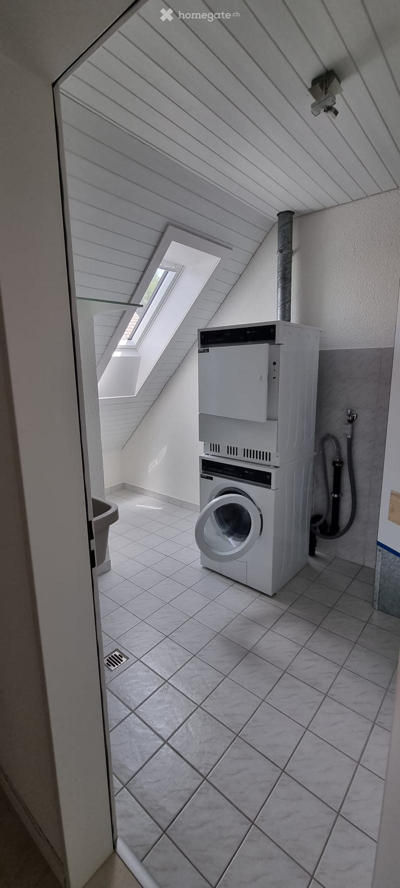 Modern bathroom with white tiled floor, white walls, and a stacked washing machine and dishwasher unit. Skylight windows provide natural light.