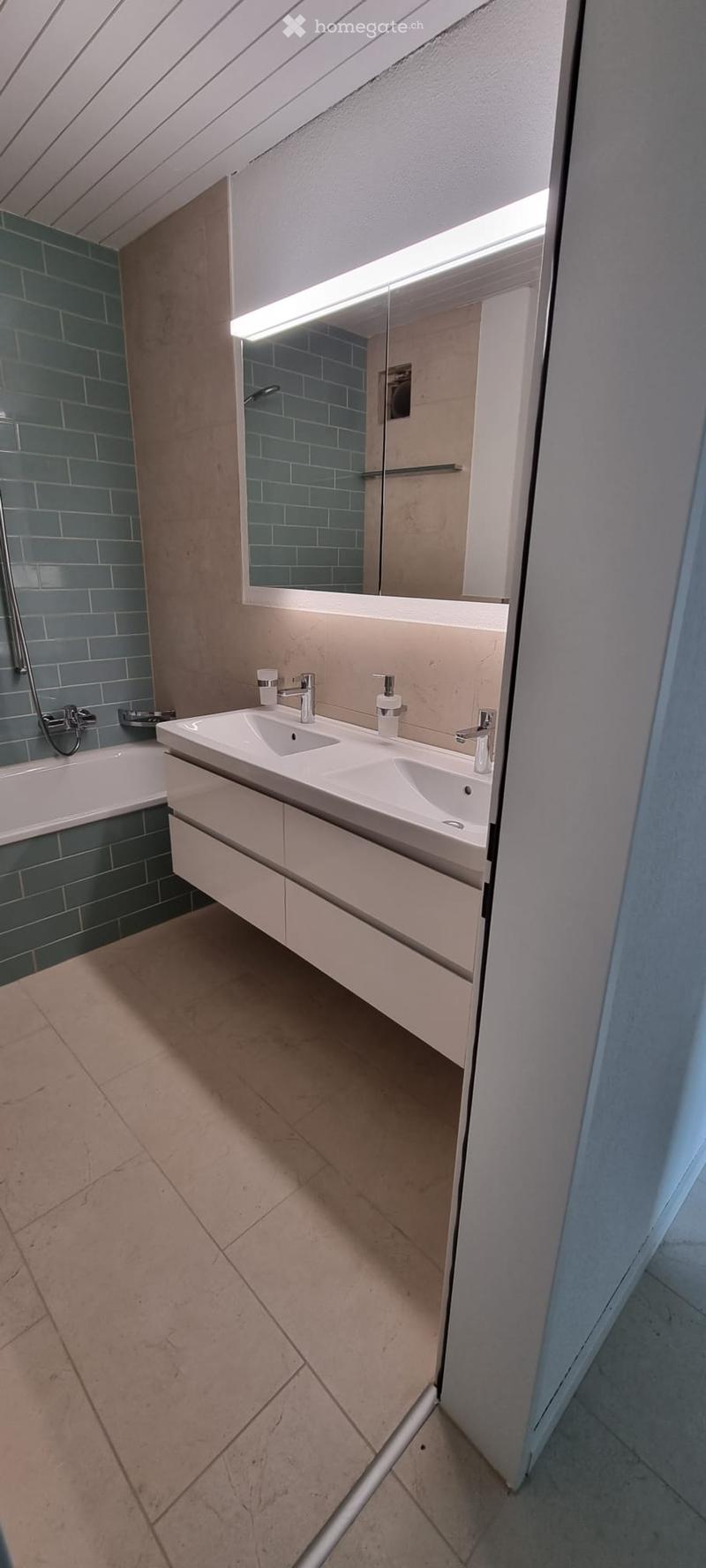 Modern bathroom with double sink, large mirror with built-in lighting, bathtub, and shower area with blue tiles.