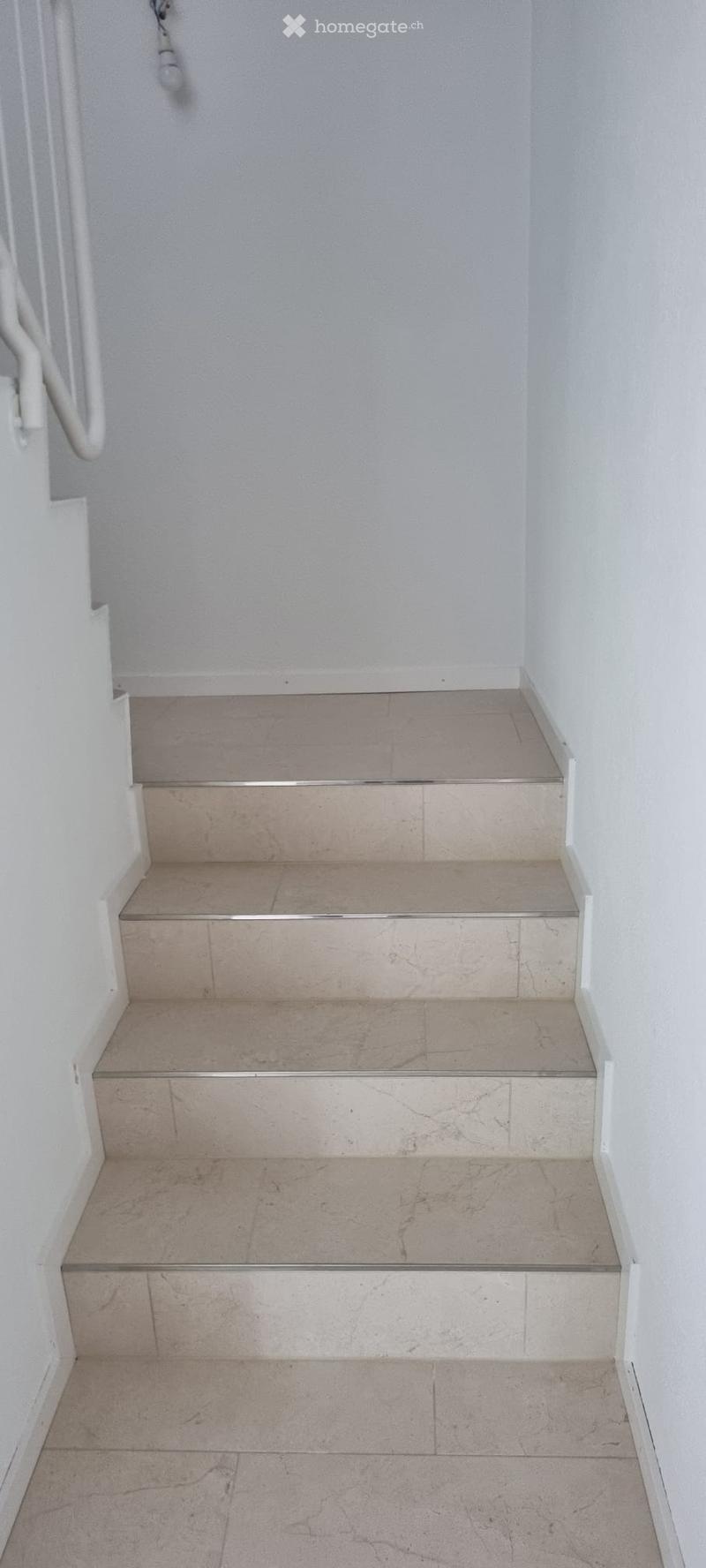 A short staircase with beige tiled steps and white walls, featuring a handrail on one side.