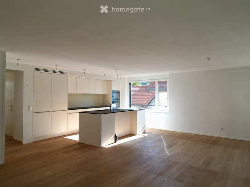 Modern kitchen with light-colored cabinetry, a central island, and wooden flooring. The space is well-lit with large windows offering a view of the roof.