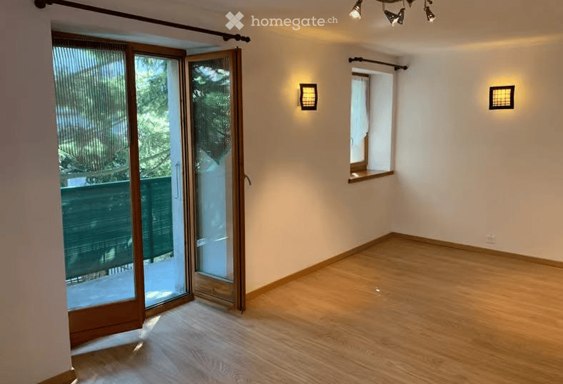 Empty room with wooden flooring, large sliding glass door leading to a balcony, two wall-mounted lights, and a small window with a white curtain.