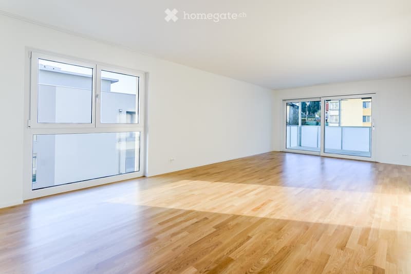 Empty room with light wooden flooring, white walls, large windows, and sliding glass doors leading to a balcony.