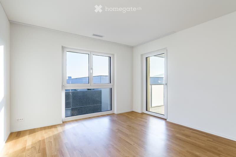 Empty room with white walls, wooden flooring, large windows, and a sliding glass door leading outside.