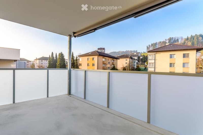 Modern balcony with white walls, grey metal railing, and a retractable awning, overlooking a residential area with trees and buildings.