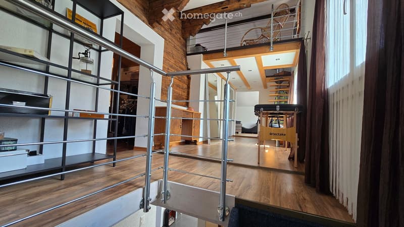 A modern, open-concept living space with wooden floors, exposed wooden beams, and a metal railing leading to an upper level. The room features built-in shelves, a wooden chair, a small table, and a curtained window area.