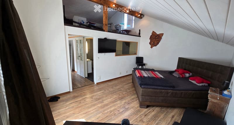 Cozy bedroom with wooden floors, a large bed with red pillows, a nightstand, a TV, a chair, and a small table in the background.