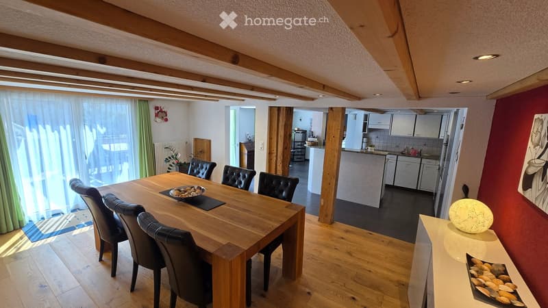 Bright dining area with wooden table and black leather chairs, adjacent modern kitchen with white cabinets, stainless steel appliances, and a small decorative lamp on a console table.
