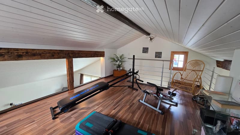 Well-lit room with wooden flooring, exercise bike, bench, wicker chair, potted plant, and a small table with books.