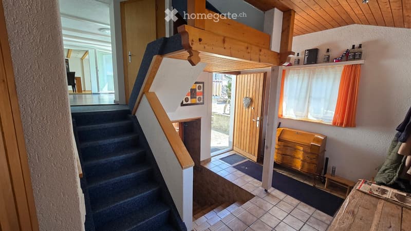 Hallway with wooden staircase, tiled floor, and a small entryway leading to another room. Features a wooden chest, a small bench, and a window with orange curtains.