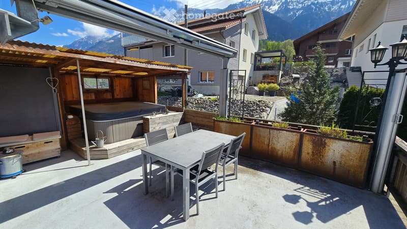 Outdoor patio with a hot tub, seating area, and dining table with chairs, surrounded by wooden structures and greenery.