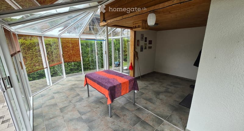 Bright conservatory with tiled floor, large windows, and a single table with a colorful tablecloth.