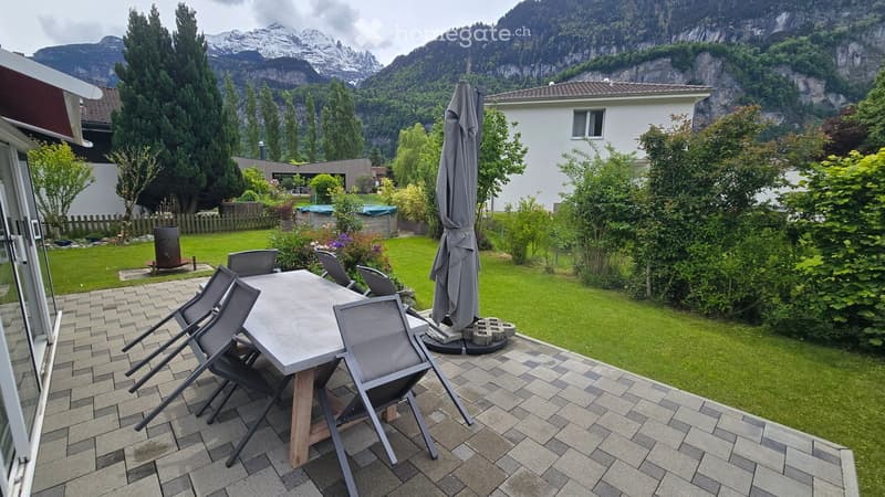 Outdoor patio with a wooden table, four chairs, and an umbrella, surrounded by a well-maintained lawn and trees, with a scenic mountain view in the background.