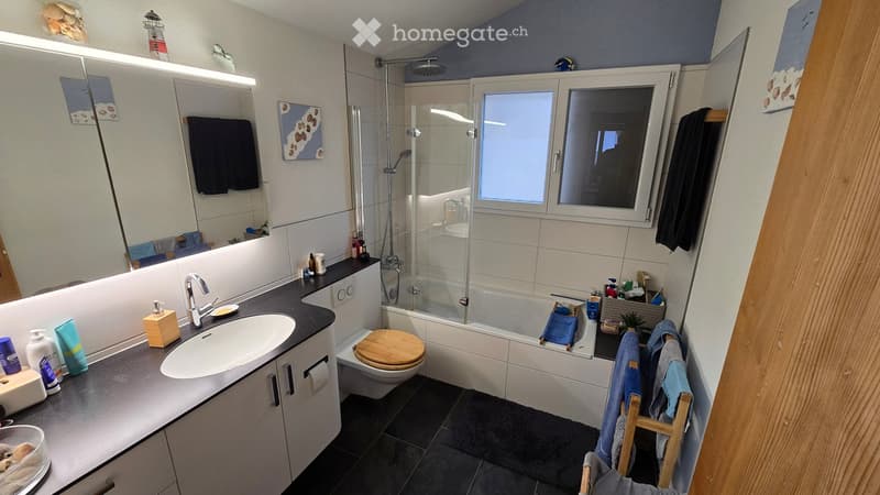Modern bathroom with a large sink, toilet, bathtub with a glass shower enclosure, and a window. The space includes a towel rack with towels, a stool, and various toiletries on the counter.