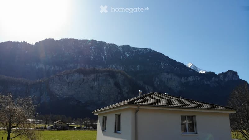 A small, single-story house with a tiled roof and white exterior, situated in a grassy field with a large, snow-capped mountain in the background.