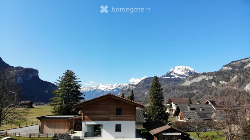 A wooden house with a sloped roof and white walls, surrounded by trees and mountains in the background.