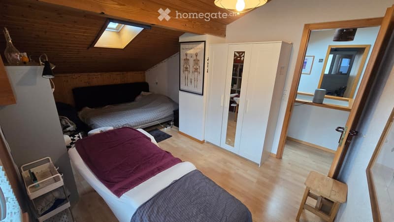 Cozy attic bedroom with wooden beams, two beds, a wardrobe, a mirror, and a small stool.