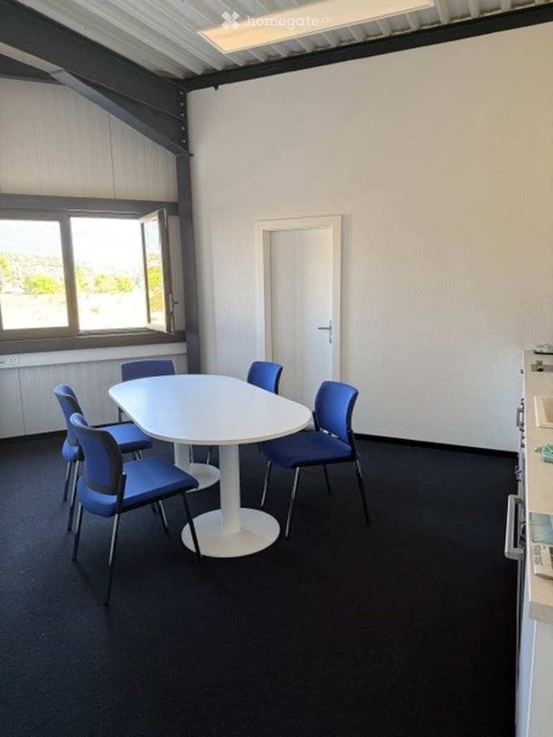 A modern, minimalistic room with a white table and blue chairs arranged around it. The room features white walls, a large window with a view, and a door leading to another space.