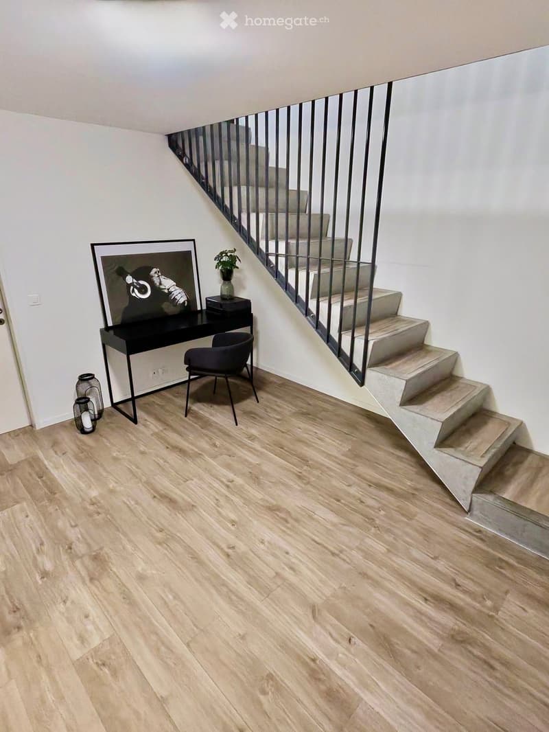 Modern hallway with wooden flooring, a staircase with metal railing, a small black table with a picture frame, a chair, and a small plant on the table.