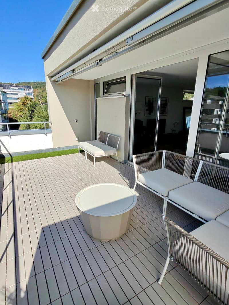 Balcony with tiled floor, white table, and two white chairs, with a view of greenery and neighboring buildings.