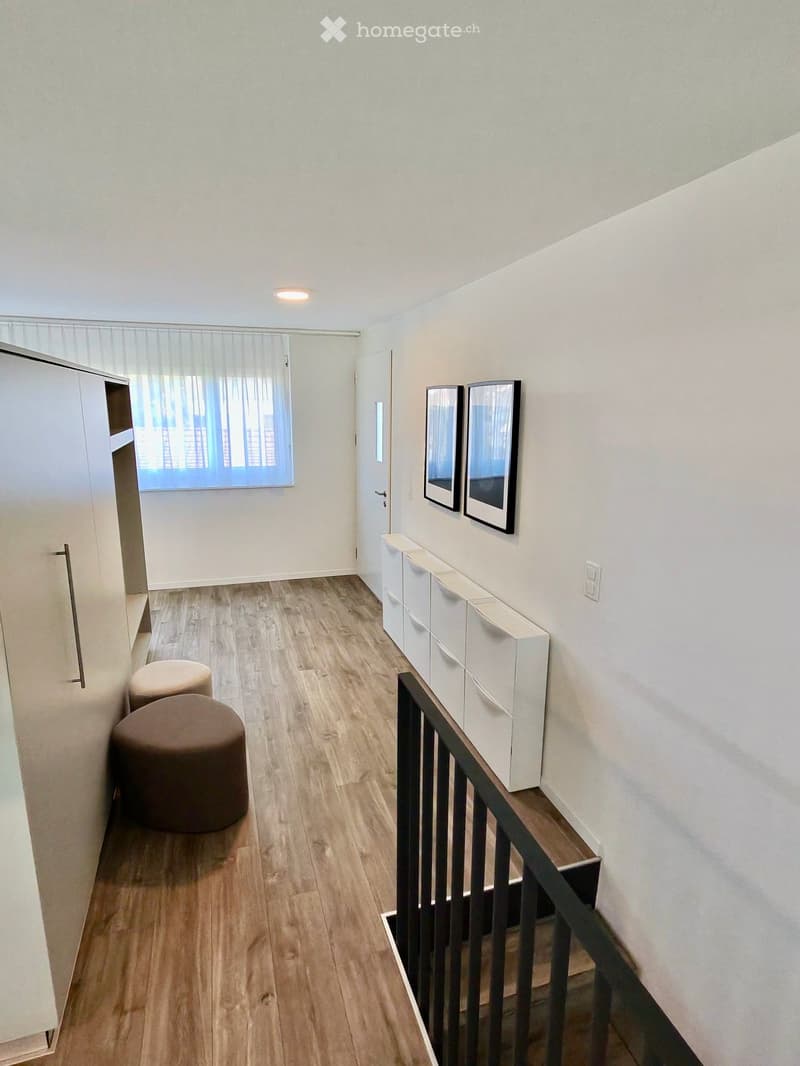A modern hallway with wooden flooring, white walls, and a staircase leading upwards. Features include built-in white storage units, two framed mirrors, a window with blinds, and two round ottomans placed near the wall.