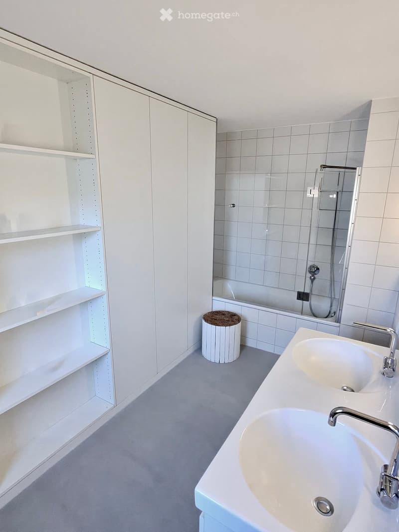 Modern bathroom with white tiles, a double sink, a bathtub, a small round stool, and built-in white cabinets with shelves.