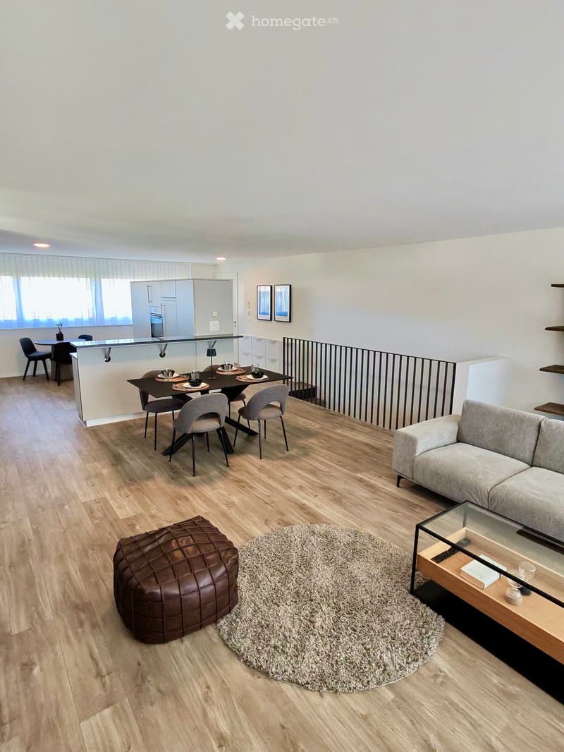 Modern living and dining area with light wood flooring, a grey sofa, a glass coffee table, a brown leather ottoman, a dining table with chairs, and a kitchen area with cabinets and a countertop.